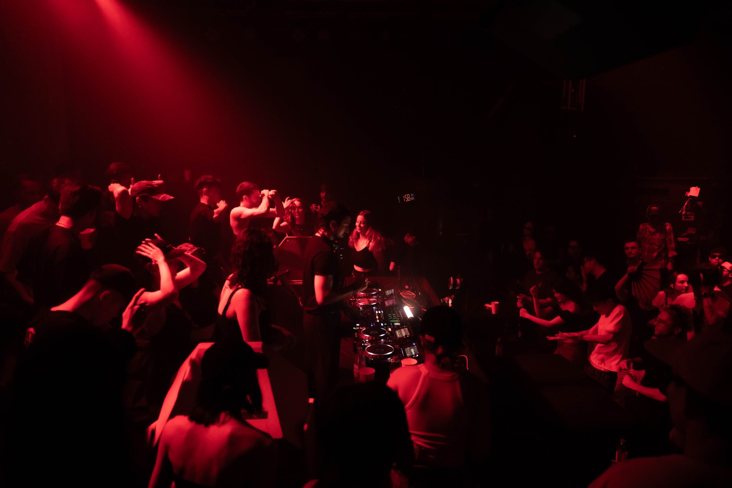 A packed floor in red light, the booth in the middle of the crowd